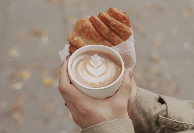 Hände halten einen Keramikbecher mit Cappuccino und Latte Art, dazu ein Franzbrötchen in Papier eingewickelt, im Hintergrund liegen herbstliche Blätter auf dem Boden.