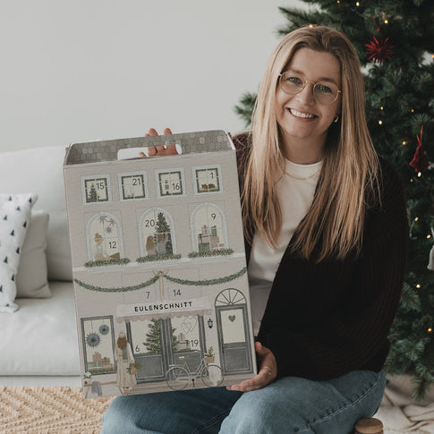 Frau hält den Eulenschnitt Signature-Adventskalender vor einem geschmückten Weihnachtsbaum.