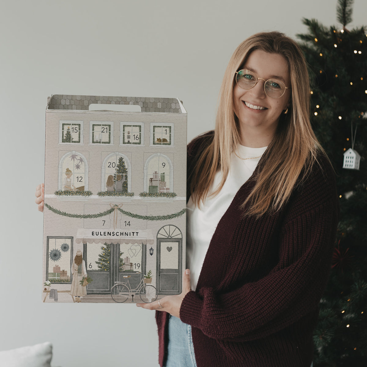 Frau präsentiert den Eulenschnitt Signature-Adventskalender neben einem beleuchteten Weihnachtsbaum.