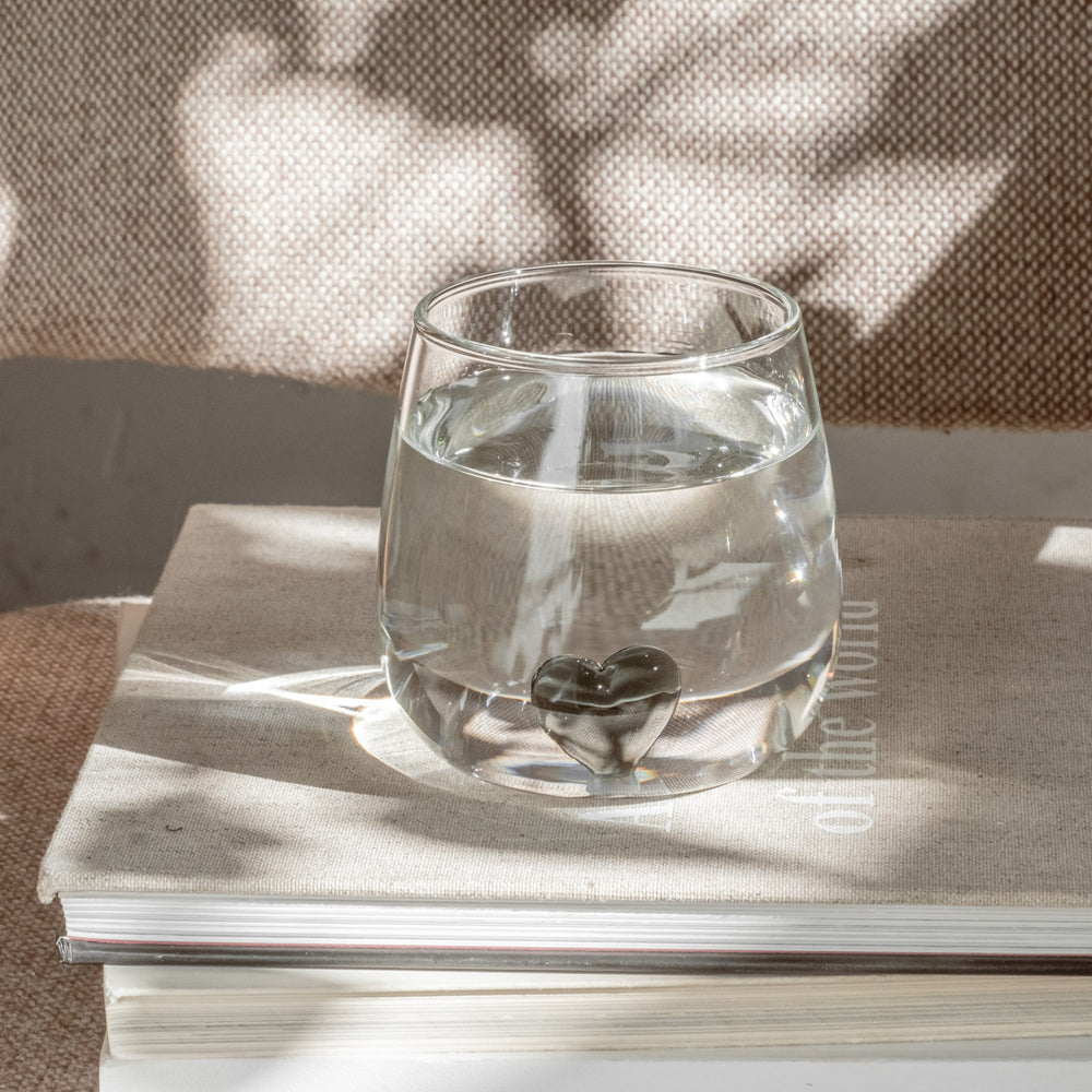 Klares Trinkglas mit schwarzer Herzfigur am Boden, gefüllt mit Wasser, auf Buch stehend.