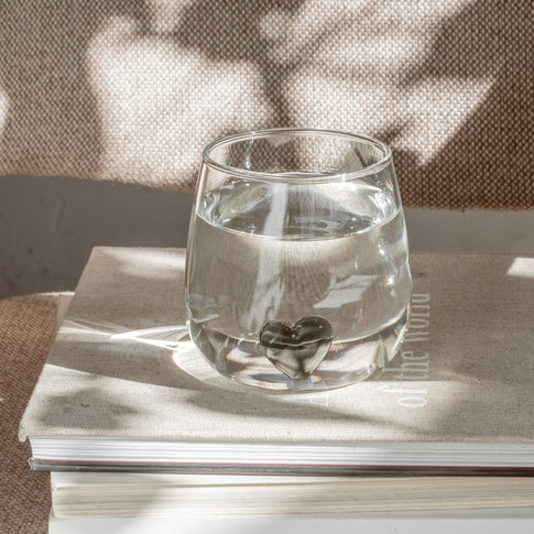 Klares Trinkglas mit schwarzer Herzfigur am Boden, gefüllt mit Wasser, auf Buch stehend.