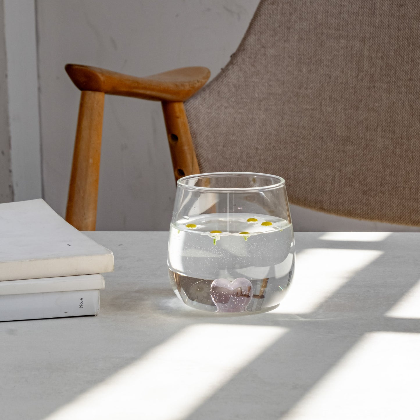 Rosa Herzfigur Trinkglas mit Wasser und kleinen Gänseblümchen gefüllt. Auf einem Tisch neben Büchern fotografiert, im Hintergrund Holzstuhl mit Stoff bezogen.