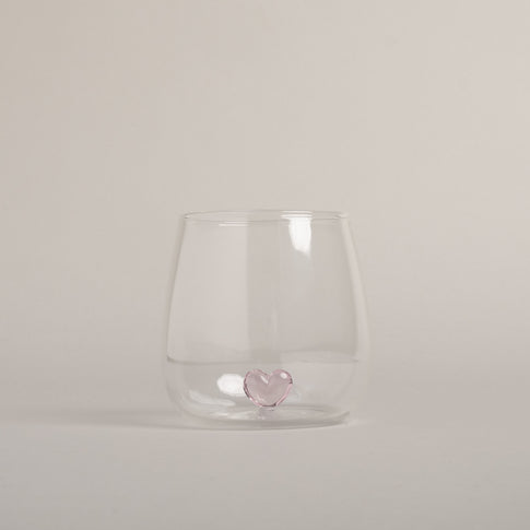 Klares Trinkglas mit rosa Herzfigur am Boden, schlichtes Design auf hellem Boden fotografiert.