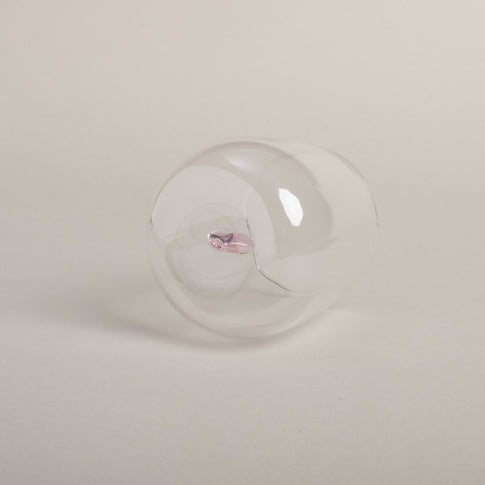 Klares Trinkglas mit rosa Herzfigur am Boden, schlichtes Design auf hellem Boden von unten fotografiert.