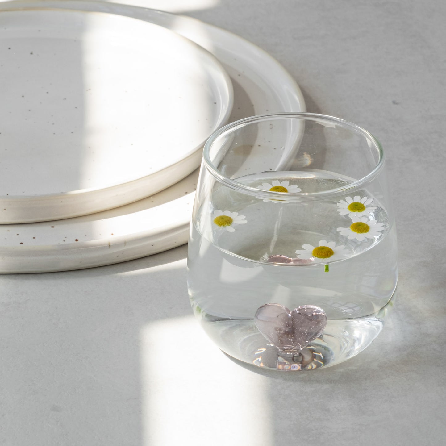 Trinkglas Herzfigur rosa mit Wasser und fünf kleinen Gänseblümchen, auf grauem Untergrund neben zwei Steingut Tellern.