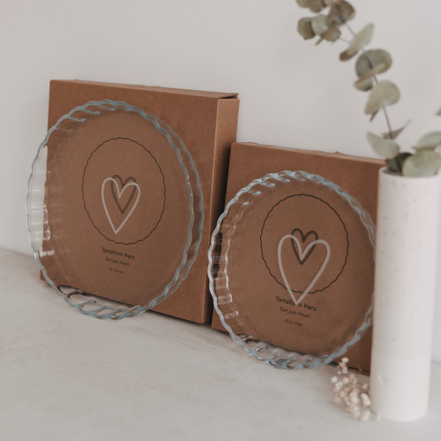Zwei Tarteformen aus Glas mit Herzprint vor brauner Verpackung nebeneinander arrangiert.