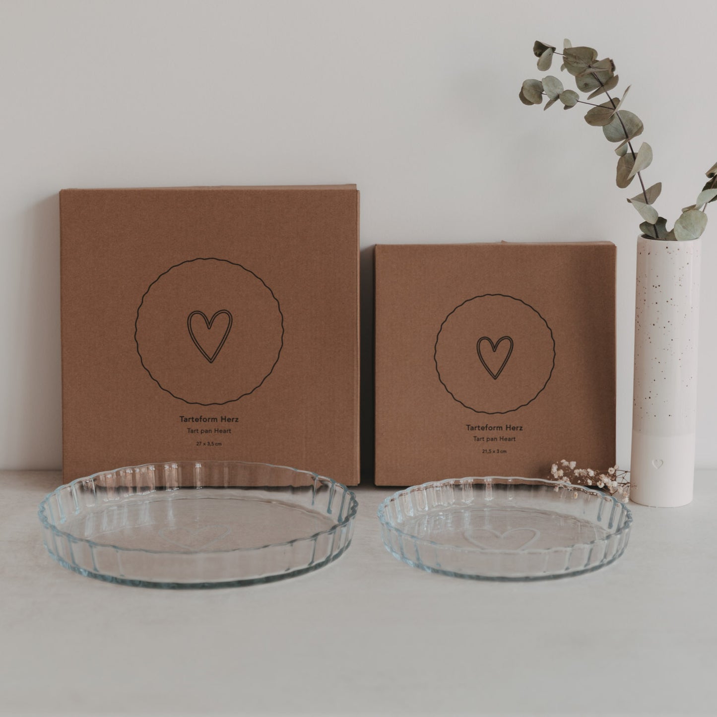 Tarteformen aus Glas in zwei Größen mit Herzdesign vor Verpackung und Vase mit Eukalyptus.