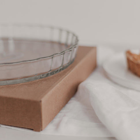 Große Tarteform Herz aus Glas auf brauner Verpackung neben Teller mit Tortenstück auf weißer Tischdecke.