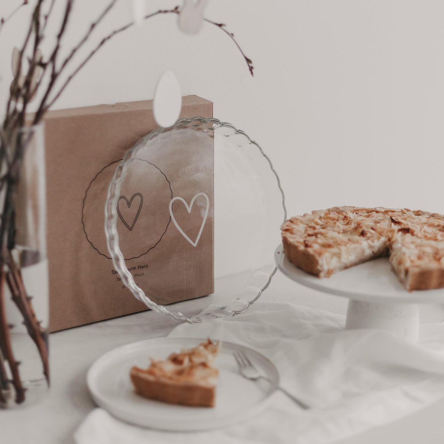Transparente Tarteform Herz aus Glas mit weißem Herzprint steht dekorativ neben angeschnittener Tarte.
