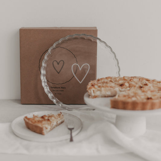 Tarteform Herz aus Glas mit Verpackung und Tarte auf Tortenplatte, vorderes Tortenstück auf Teller serviert.
