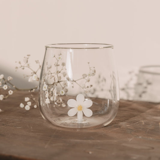 Figurglas Daisy mit einer weißen Blume im Glas, Trinkglas auf Holztisch mit zarten weißen Blüten im Hintergrund