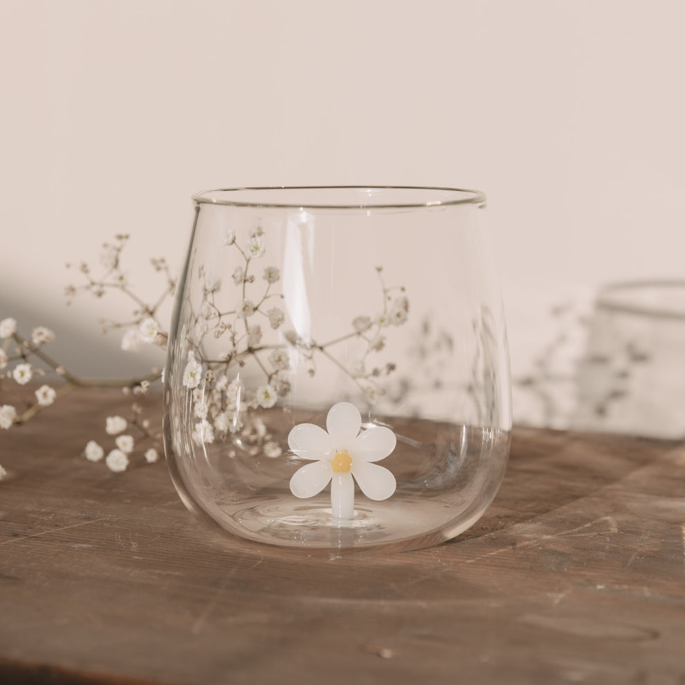 Figurglas Daisy mit einer weißen Blume im Glas, Trinkglas auf Holztisch mit zarten weißen Blüten im Hintergrund