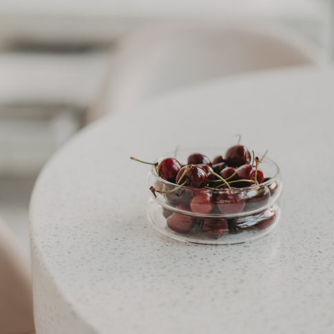 Gefüllt mit Kirschen (Tisch) – Runde Glas-Bubbles-Schale klein mit roten Kirschen auf hellem Tisch dekoriert.