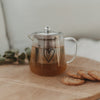 Teekanne aus Glas mit schwarzem Herzmotiv, gefüllt mit Tee, auf Holztablett mit Keksen und Eukalyptuszweig