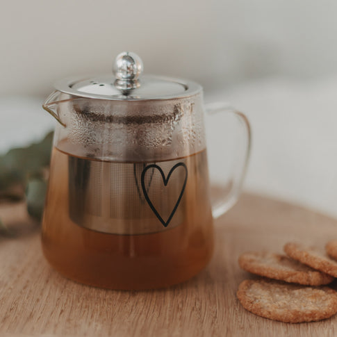 Glasteekanne mit Teesieb und schwarzem Herz, dampfend heiß, auf Holzunterlage mit Keksen serviert