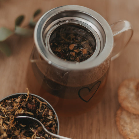 Offene Teekanne mit losem Tee im Edelstahlsieb, neben einer Dose mit Kräutertee und Cookies
