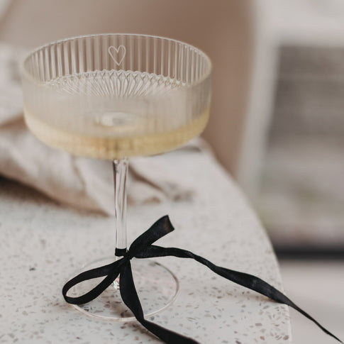 Champagnerglas mit Herzgravur und schwarzer Schleife steht auf hellem Tisch.
