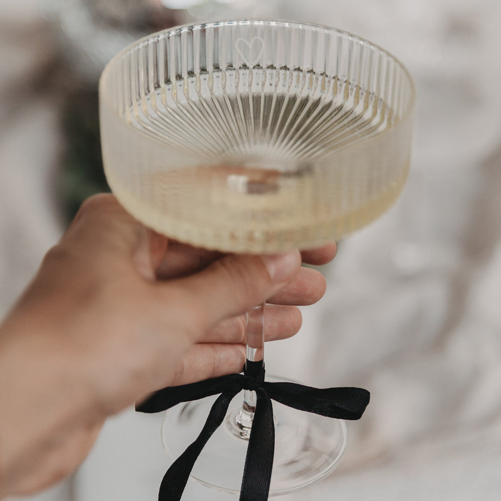Hand hält elegantes Champagnerglas mit Herzgravur und schwarzer Schleife am Stiel.