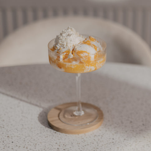 Elegantes Champagnerglas mit Dessert und Sahnehaube auf Holzuntersetzer, stilvoll auf hellem Tisch platziert.