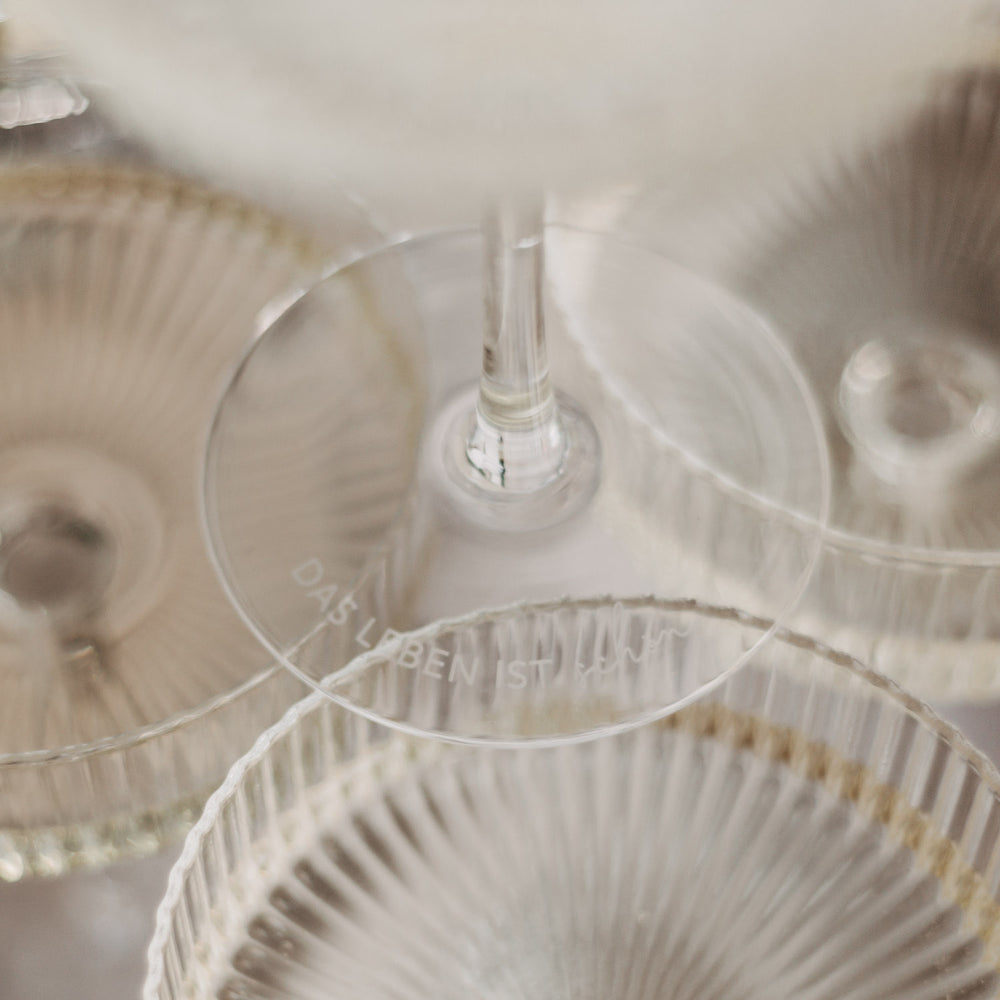 Detailansicht der Gravur „Das Leben ist schön“ auf dem Fuß eines Glases mit weiteren Gläsern im Hintergrund.