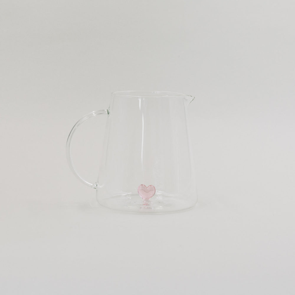 Karaffe aus Glas mit rosa Herzfigur im Inneren, minimalistisch vor weißem Hintergrund inszeniert.