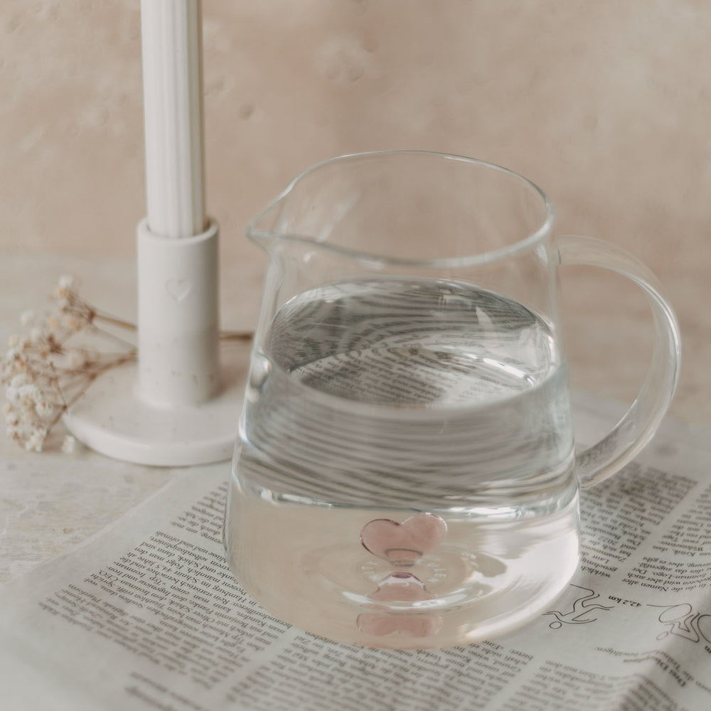 Karaffe mit Wasser und rosa Herz auf Zeitung, neben Kerzenständer mit Herzdetail.