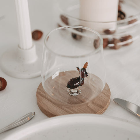 Trinkglas mit brauner 3D-Rehfigur auf einem Holzuntersetzer, dekorativ gedeckt mit Nüssen und Kerzen.