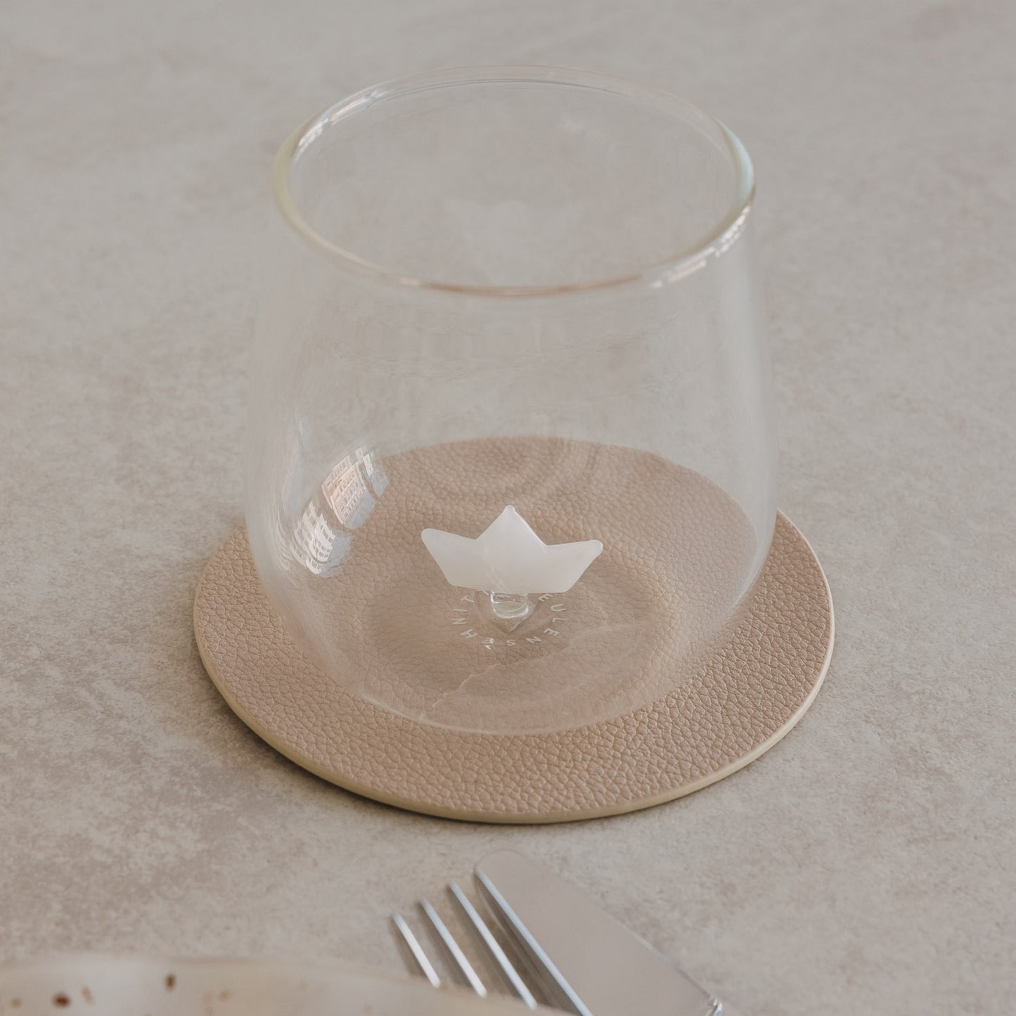 Trinkglas mit Papierboot Motiv auf Untersetzer, modernes Glas neben Teller und Besteck