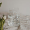 Glasvase mit Blumenwiesen-Motiv gefüllt mit Wasser auf hellem Stoff mit Tulpe daneben