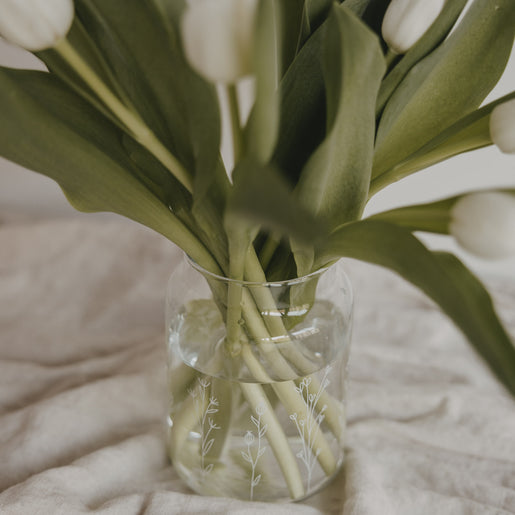 Glasvase mit Blumenwiesen-Motiv und weißen Tulpen, dekorativ auf Leinendecke