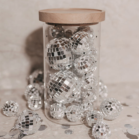 Hoher Glasbehälter mit kleinen schwarzen Herzen und Holzdeckel, gefüllt mit silbernen Discokugeln und umgeben von weiteren Discokugeln und Konfetti.