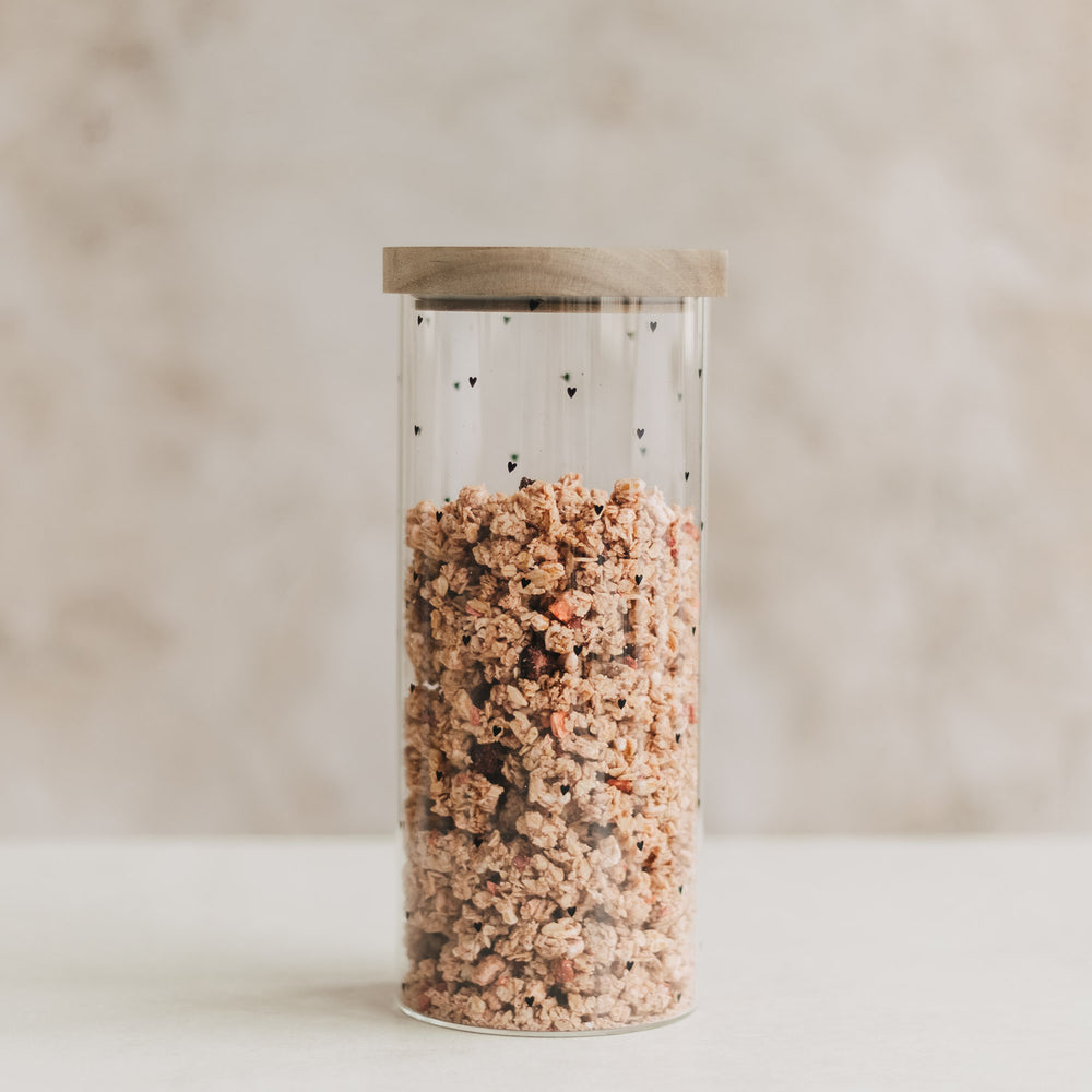 Hohes Vorratsglas mit kleinen schwarzen Herzen, Holzdeckel und Müsli auf hellem Hintergrund.
