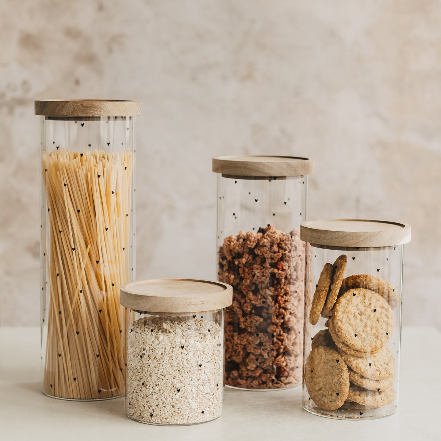 Vier Vorratsgläser mit schwarzen Herzen und Holzdeckeln, gefüllt mit Spaghetti, Haferflocken, Müsli und Keksen.