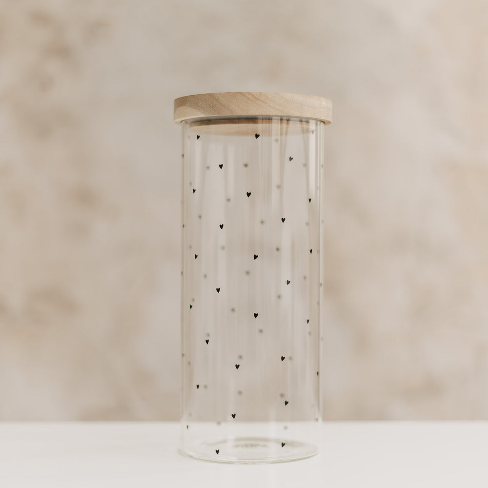 Leeres hohes Vorratsglas mit kleinen schwarzen Herzen und hellem Holzdeckel auf neutralem Hintergrund.