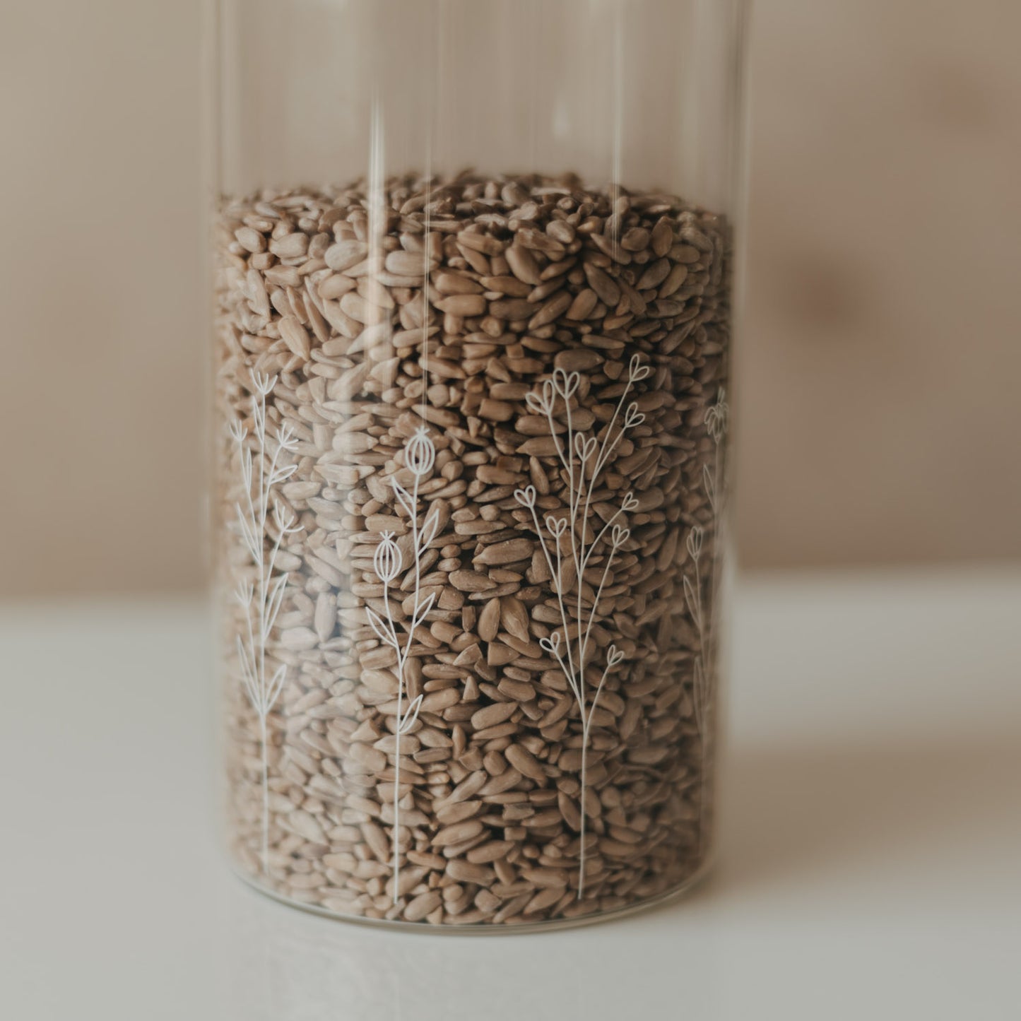 Detailansicht eines Glas-Vorratsbehälters mit filigranem weißem Wildblumen-Motiv und Sonnenblumenkernen