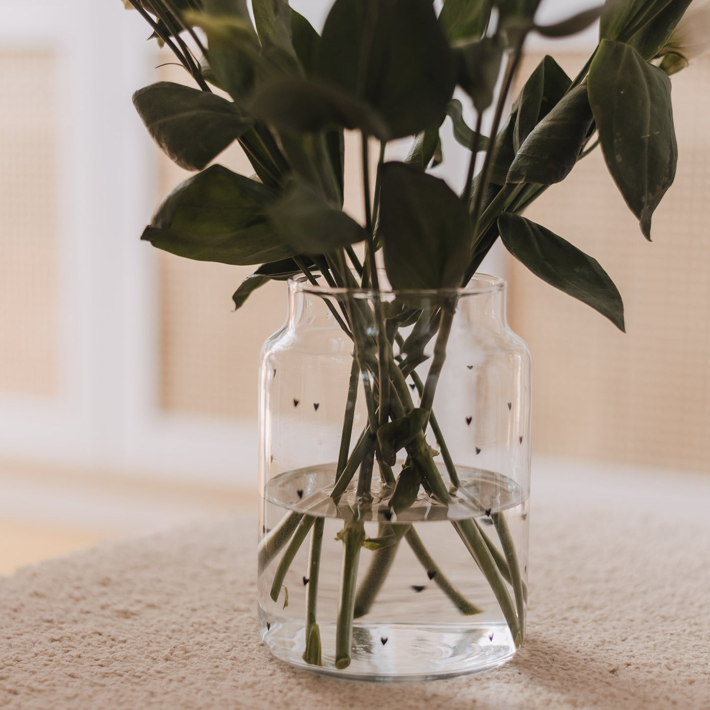 Glasvase mit weißen Herzen und Blumen auf Stoffhocker