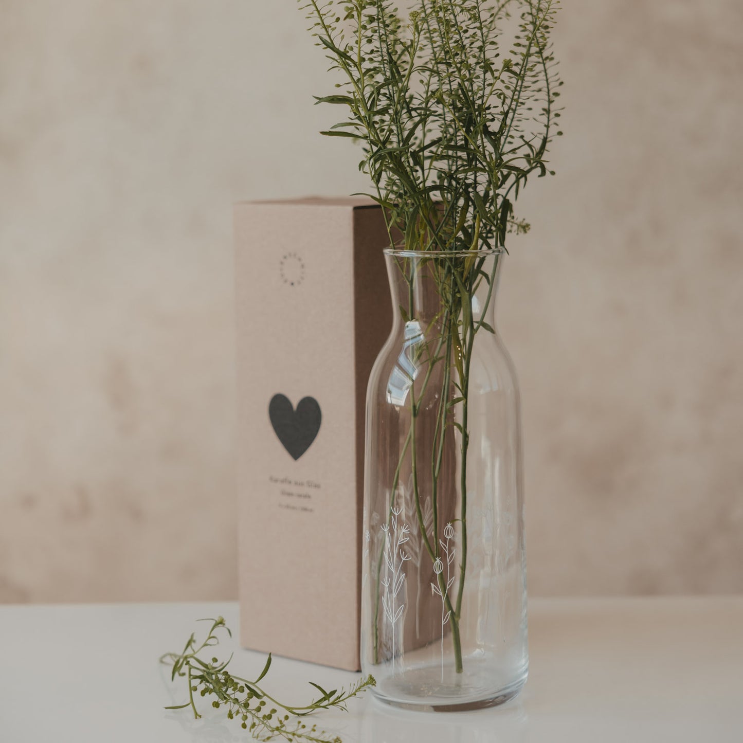 Glas Karaffe mit Blumenmotiv und frischen Zweigen, neben Verpackung mit Herzmotiv