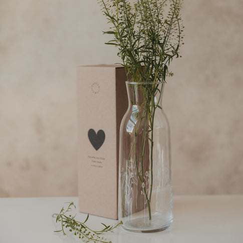 Glas Karaffe mit Blumenmotiv und frischen Zweigen, neben Verpackung mit Herzmotiv