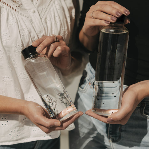 Zwei Glas Trinkflaschen im Vergleich, eine mit Herzmotiv, eine mit floralem Muster, in Händen gehalten