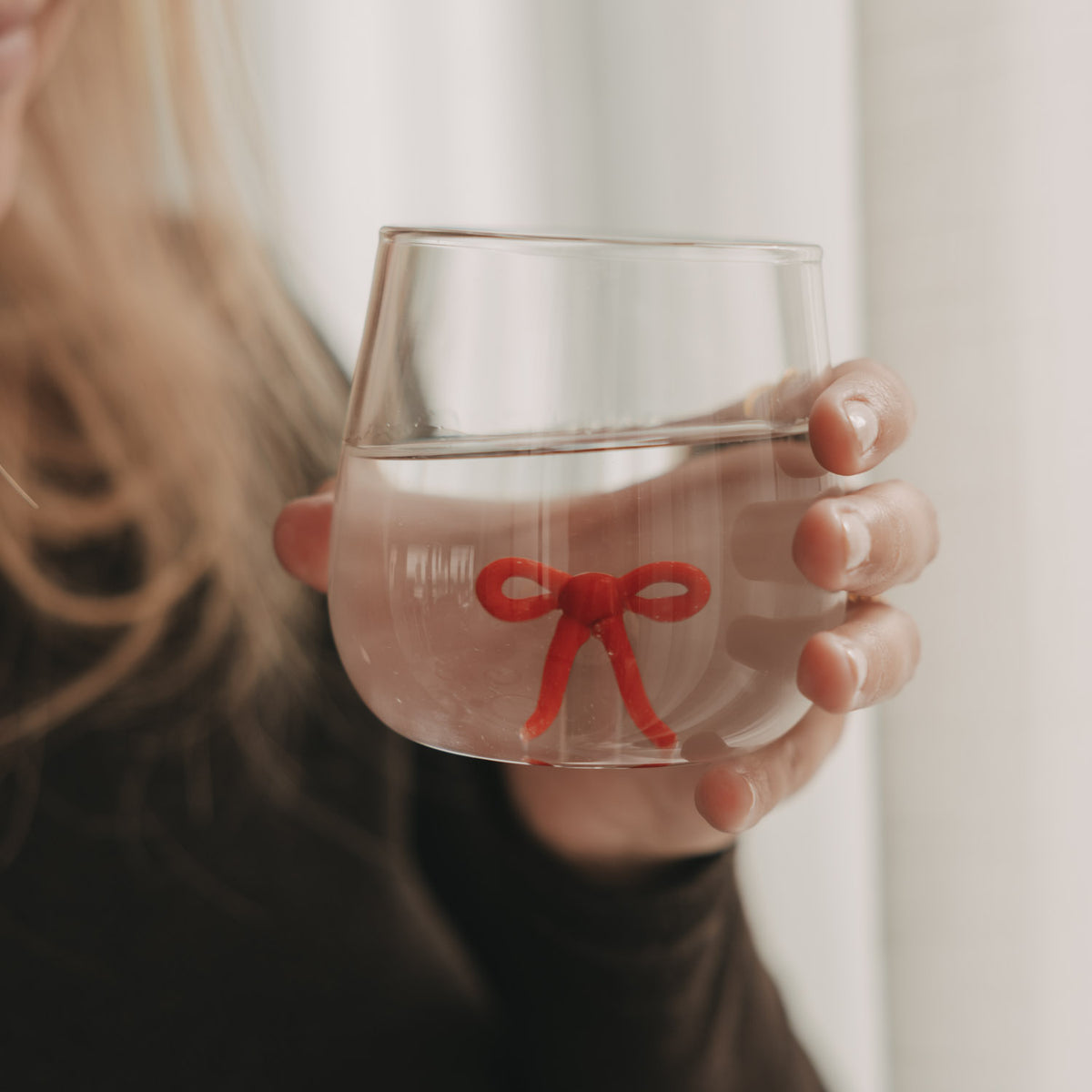 Frau hält ein mit Wasser gefülltes Figurenglas mit roter Schleife im Glasboden vor hellem Hintergrund.