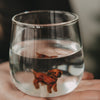 Nahaufnahme eines Wasserglases mit brauner Dackelfigur im Glasboden, aus leicht erhöhter Perspektive.