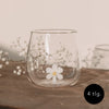 Figurglas Daisy mit einer weißen Blume im Glas, Trinkglas auf Holztisch mit zarten weißen Blüten im Hintergrund
