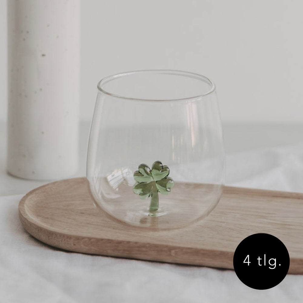 Trinkglas mit einer grünen Kleeblattfigur im Glasboden, auf einem Holztablett platziert, heller Hintergrund.