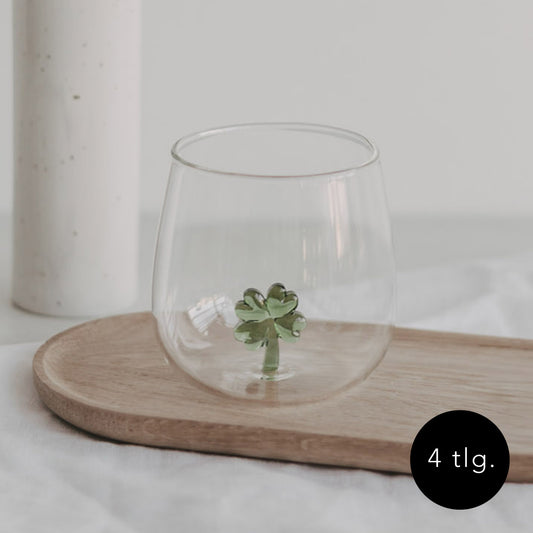 Trinkglas mit einer grünen Kleeblattfigur im Glasboden, auf einem Holztablett platziert, heller Hintergrund.