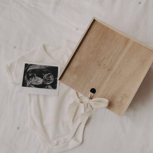 Holz-Erinnerungsbox mit Gänsegravur neben Babybody und Ultraschallfoto auf weißer Decke.