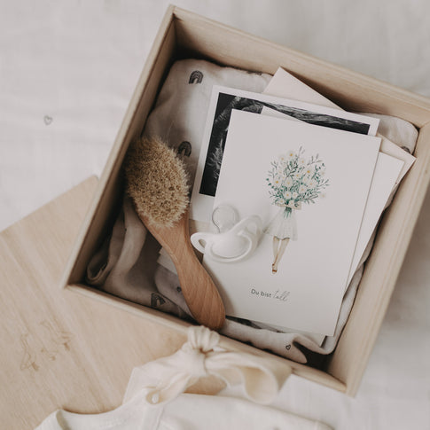 Geöffnete Erinnerungsbox gefüllt mit Babyartikeln wie Karte, Schnuller, Bürste und Tuch mit Regenbogenmotiv.