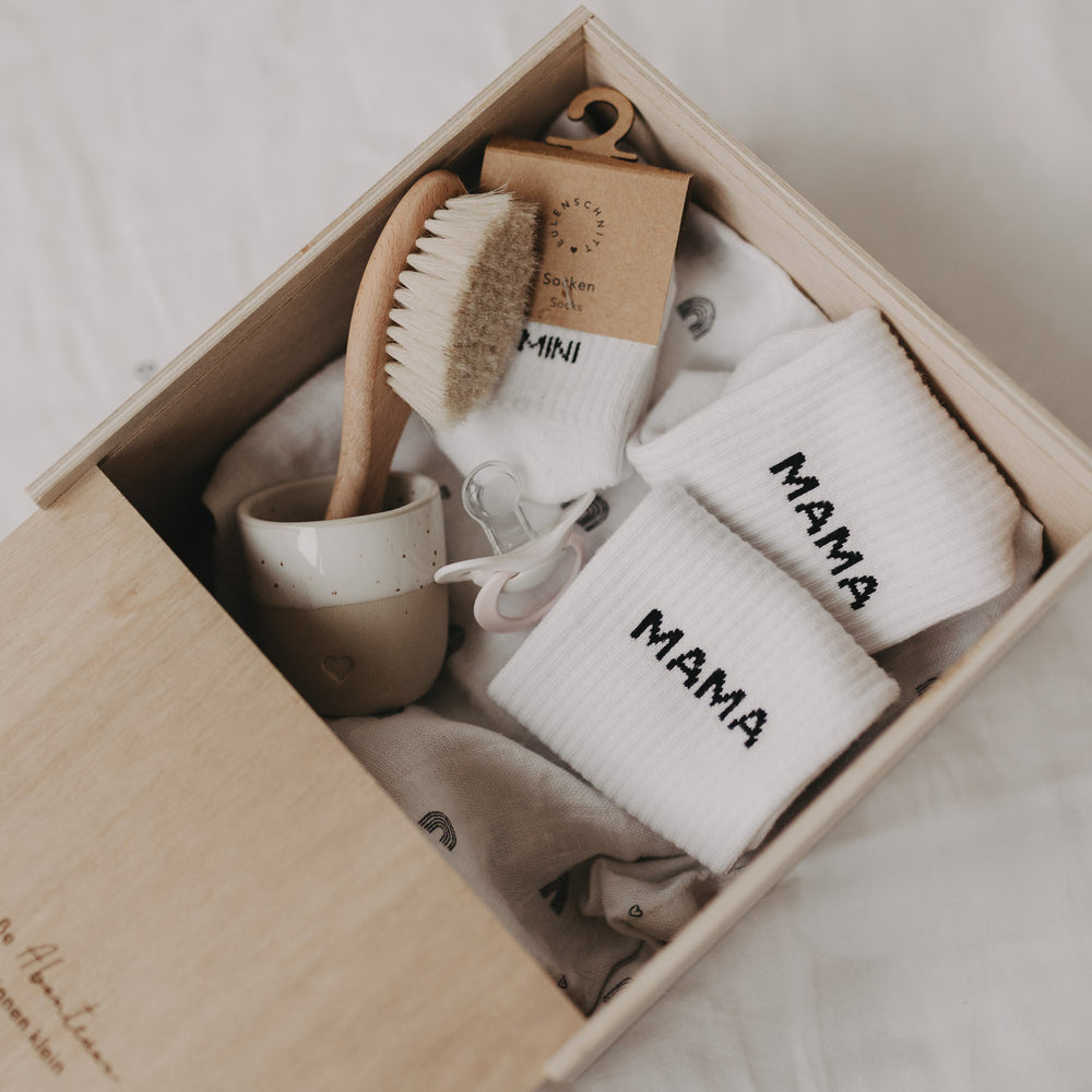 Geöffnete Erinnerungsbox aus Holz, gefüllt mit Babyartikeln wie Mini-Söckchen, Bürste und Schnuller.