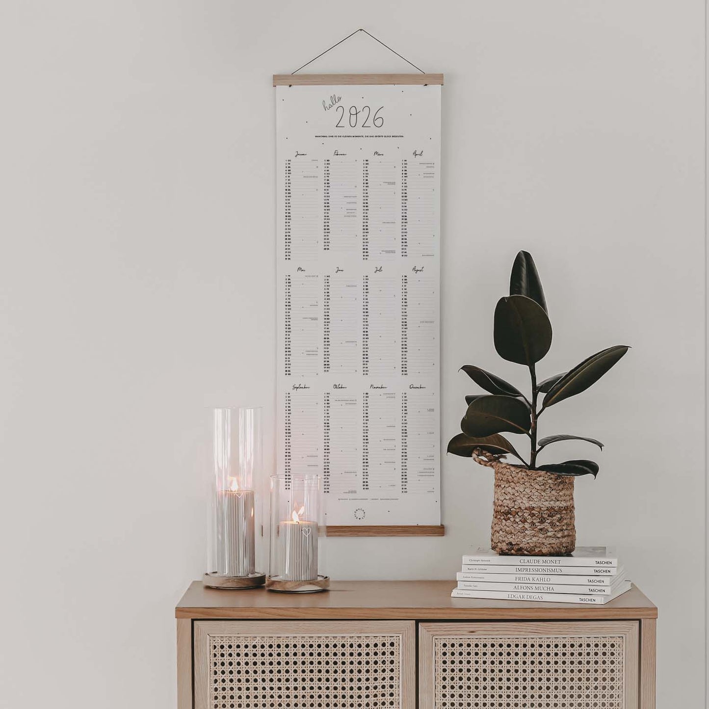Wandkalender 2026 im Hochformat mit Holzleiste, dekorativ über Kommode mit Kerzen und Pflanze aufgehängt.