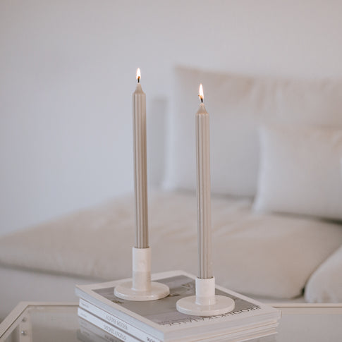 Zwei graue Stabkerzen in weißen Kerzenhaltern auf einem Glastisch vor heller Couch im Hintergrund