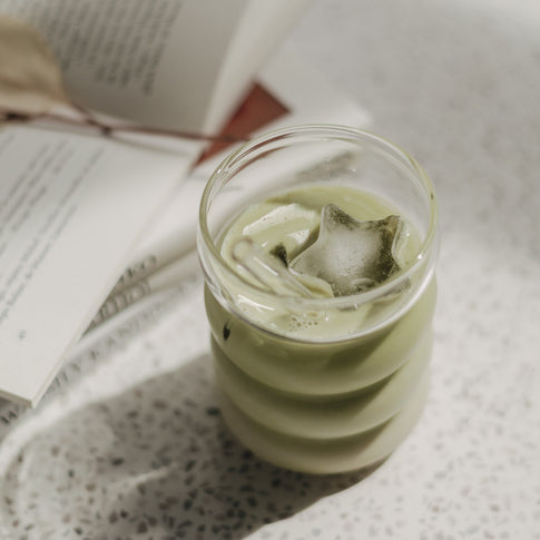 Glas mit grünem Matcha und sternförmigem Eiswürfel auf hellem Tisch, dekorativ inszeniert mit offenem Buch im Hintergrund.